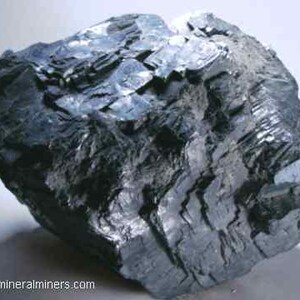 May include: A dark gray mineral specimen with a layered, crystalline structure. The surface reflects light, showing a metallic sheen. The rough, irregular shape suggests a natural, unpolished state. The image is of a mineral sample.
