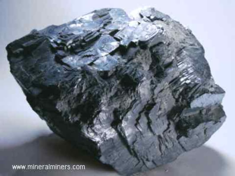 May include: A dark gray mineral specimen with a layered, crystalline structure. The surface reflects light, showing a metallic sheen. The rough, irregular shape suggests a natural, unpolished state. The image is of a mineral sample.