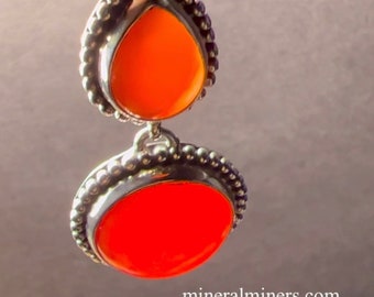 May include: A pair of dangle earrings with two orange gemstones set in silver. The top gemstone is teardrop shaped and the bottom gemstone is round. The earrings have a beaded border.