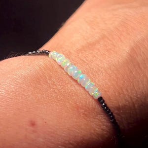 May include: A delicate bracelet featuring a strand of iridescent opal beads interspersed with black beads. The bracelet is worn on a wrist, with the text "MineralMiners.com" visible.