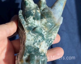 Puede incluir: Una escultura de cabeza de dragón tallada en tonos verdes, blancos y marrones. Los cuernos del dragón son puntiagudos y los detalles de la cara son visibles. La escultura se sostiene en una mano sobre un fondo azul. El sitio web "MineralMiners.com" es visible.