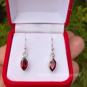 May include: A pair of silver dangle earrings with red gemstone drops. The earrings are in a red velvet box.