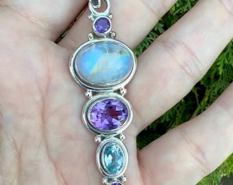 May include: A silver pendant with a rainbow moonstone and amethyst gemstones. The pendant is on a hand with a green background.