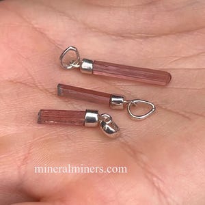 May include: Three pink tourmaline crystal pendants with silver bail settings. The pendants are all different sizes and shapes. The crystals are a light pink color and have a smooth, polished finish.