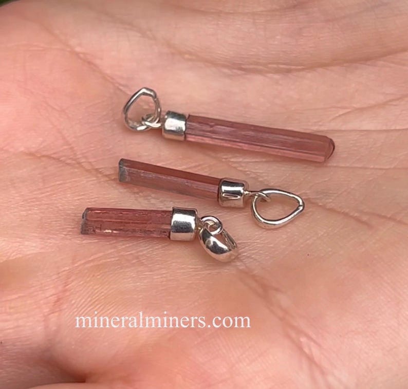 May include: Three pink tourmaline crystal pendants with silver bail settings. The pendants are all different sizes and shapes. The crystals are a light pink color and have a smooth, polished finish.