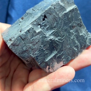 May include: A large, dark gray mineral specimen with a cubic shape and a rough, textured surface. The mineral is held in a hand against a blue background. The website mineralminers.com is visible in the bottom right corner.