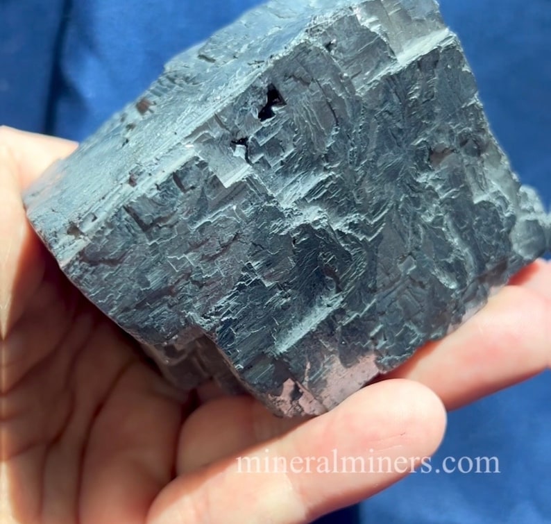 May include: A large, dark gray mineral specimen with a cubic shape and a rough, textured surface. The mineral is held in a hand against a blue background. The website mineralminers.com is visible in the bottom right corner.