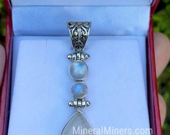May include: A silver pendant with three moonstone gemstones. The top has an ornate design, followed by two round stones, and a teardrop-shaped stone at the bottom. The pendant is displayed in a red jewelry box.