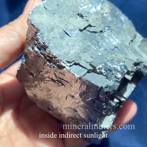 May include: A close-up of a mineral specimen with a metallic sheen. The rock has a layered, fractured appearance with shades of gray, silver, and brown. The text "mineralminers.com inside indirect sunlight" is visible.