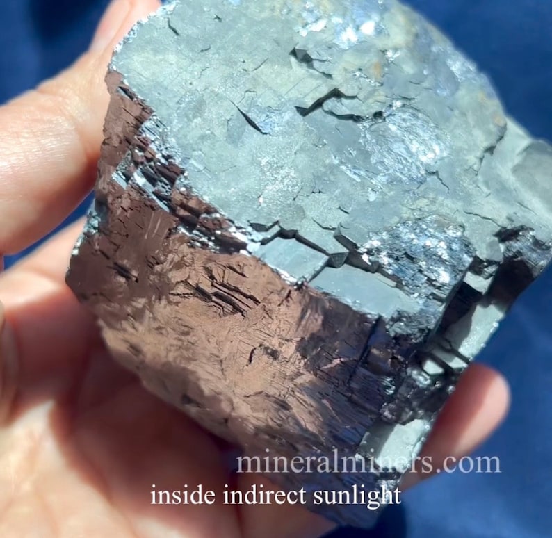 May include: A close-up of a mineral specimen with a metallic sheen. The rock has a layered, fractured appearance with shades of gray, silver, and brown. The text "mineralminers.com inside indirect sunlight" is visible.