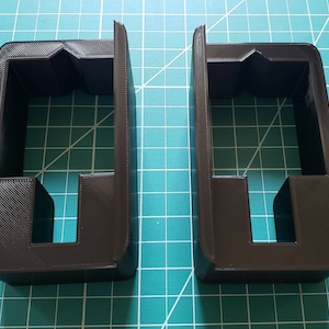 May include: Two dark brown, rectangular 3D-printed objects with a unique design. Each has a tall, open-top frame with a smaller, rectangular base and a central cutout. The objects are placed on a green grid surface.