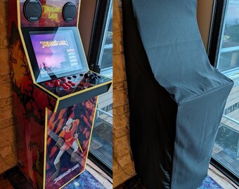 May include: An arcade cabinet featuring the game "Dragon's Lair." The cabinet has a red and gold design with a screen, joystick, and buttons. A black cover is on the right. The cabinet has speakers and the game's logo.