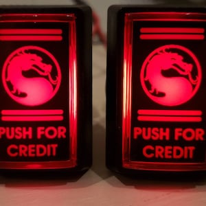 May include: Two rectangular black buttons with a red illuminated design. Each button features a red dragon logo within a circle, with the text "PUSH FOR CREDIT" below. The buttons are lit with a bright red glow.