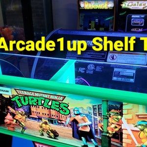May include: A green Arcade1up shelf topper with the Teenage Mutant Ninja Turtles logo. The shelf topper has a neon green trim and the text "Arcade1up Shelf Topper!" is displayed. Other arcade game titles are visible in the background.
