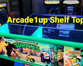 Könnte beinhalten: Ein grüner Arcade1up-Regalaufsatz mit dem Logo der Teenage Mutant Ninja Turtles. Der Regalaufsatz hat eine neongrüne Umrandung und der Text "Arcade1up Shelf Topper!" ist zu sehen. Weitere Arcade-Spieletitel sind im Hintergrund sichtbar.