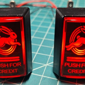 May include: Two rectangular arcade push buttons with black plastic housings and translucent red covers. Each button features a graphic of a running rabbit inside a circular ring and the text PUSH FOR CREDIT printed in red on the lower section.