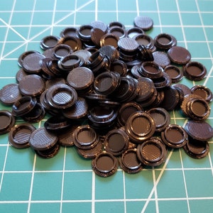 May include: A pile of small, dark brown plastic discs with a circular design. The discs have a raised, textured surface and a recessed center. The objects are scattered on a green grid surface.