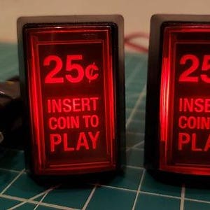 May include: Two rectangular arcade-style push buttons with black frames and illuminated red translucent covers. Each button features the white text 25¢ INSERT COIN TO PLAY. A black USB cable with yellow and black wiring is attached to the left button.