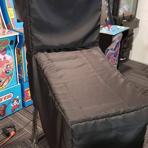 May include: A black pinball machine cover. The cover is made of a dark, possibly nylon, fabric and is draped over the pinball machine. The legs of the pinball machine are chrome. Other arcade games are visible in the background.