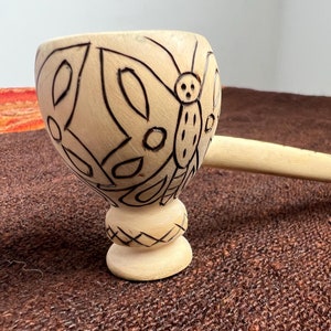 May include: A wooden cup with a long handle, carved with a butterfly design. The cup is light brown and the butterfly is dark brown.