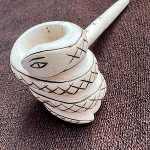 May include: A white wooden pipe carved in the shape of a coiled snake. The snake has a detailed eye and scales, and the pipe has a long, thin stem.