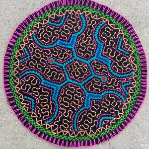 Puede incluir: Una pieza de tela negra circular con intrincados bordados hechos a mano en colores vibrantes. El diseño incluye patrones de hilo rosa neón, verde brillante, naranja y turquesa que forman formas geométricas y dos pequeños puntos rojos.