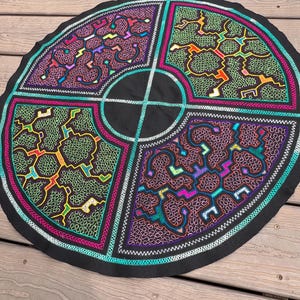 Op de afbeelding: Een rond zwart stuk stof met vier afzonderlijke kwadranten van ingewikkeld, kleurrijk borduurwerk. Elk gedeelte toont complexe, doolhofachtige patronen in levendige neonkleuren zoals groen, roze, oranje en paars, gescheiden door witte lijnen.