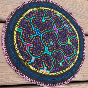 Puede incluir: Un parche circular de tela negra con un intrincado bordado multicolor en tonos amarillo, azul, naranja, rosa y turquesa. El diseño consiste en complejos patrones geométricos entrelazados tipo laberinto, con un borde cosido en zigzag rosa.
