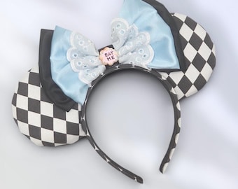 Puede incluir: Una diadema con dos orejas redondas de patrón a cuadros blanco y negro. Un lazo grande se sitúa entre las orejas, hecho de satén azul claro y borde de encaje blanco. Un pequeño dije circular rosa con el texto EAT ME está en el centro.