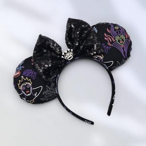May include: A headband featuring two round ears covered in black fabric with a neon-style print of various villainous characters. A large black sequined bow sits between the ears, accented with a small silver-toned crown charm with clear crystals.