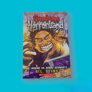 Puede incluir: Una portada de libro de Goosebumps HorrorLand con un personaje de dibujos animados con una amplia sonrisa de dientes. El título del libro es "Dr. Maniac vs. Robby Schwartz" de R.L. Stine. El libro está publicado por Scholastic.