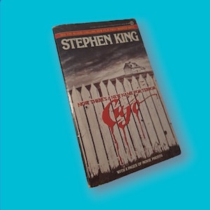 Puede incluir: Una portada de libro de bolsillo que presenta una valla blanca de madera con sangre roja goteando por los postes de la valla. El título del libro es "Stephen King" y el subtítulo del libro es "Now there's a new name for terror: " con la palabra "C.Y.A." en letras rojas. La portada del libro también incluye el texto "With 8 pages of movie photos."
