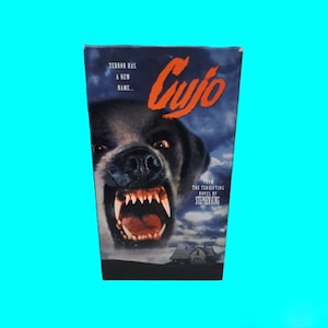 Puede incluir: Una carátula de cinta VHS para la película Cujo, basada en la novela de Stephen King. La carátula presenta un primer plano de un perro gruñendo con una expresión amenazante. El texto "Cujo" está en letras rojas grandes, y el lema "Terror has a new name..." está en letras blancas más pequeñas.