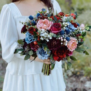 May include: A bridal bouquet with a mix of burgundy, blush pink, and blue flowers. The bouquet includes roses, peonies, and eucalyptus.
