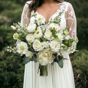 May include: A white bridal bouquet featuring a variety of white flowers, such as roses, peonies, and ranunculus. The bouquet is held by a person wearing a white dress.