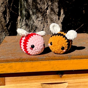 Pode incluir: Dois bichinhos de pelúcia de abelha feitos à mão em crochê. Uma abelha é rosa e vermelha, a outra é amarela, preta e branca. Ambas têm asas brancas e olhos pretos. Os bichinhos de pelúcia estão em uma superfície de madeira, com um tronco de árvore ao fundo.