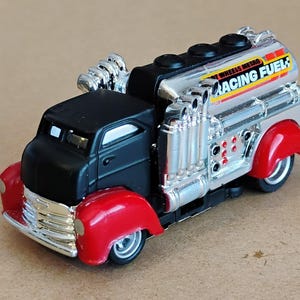 May include: A toy truck with a black cab and red fenders. The silver tank has the words "RACING FUEL" and "WHEELS METRO" printed on it. Chrome exhaust pipes and a chrome grill complete the look.