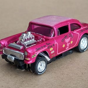 May include: Pink 1955 Chevrolet with a custom paint job and a large engine. The car has a "Stinky Kitty" decal on the side and the text "Listen to me purr - 427 cu. in." The car is a slot car.