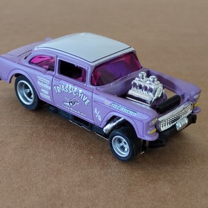 May include: Purple 1/24 scale slot car model of a 1955 Chevrolet with a white roof and a chrome engine. The car has the words "Triassic-Five" and "Fuel Injected" on the side.