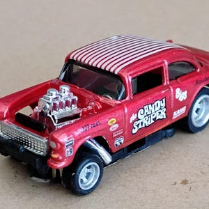 May include: A red toy car with a white striped roof and the words "the Candy Striper" on the side. The car has a chrome engine and wheels. The car is a classic model.