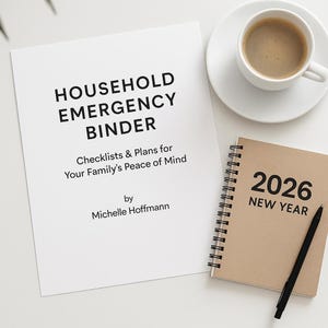 May include: A white binder titled "HOUSEHOLD EMERGENCY BINDER" with the subtitle "Checklists & Plans for Your Family's Peace of Mind" and author Michelle Hoffmann. A tan spiral-bound notebook with "2026 NEW YEAR" and a cup of coffee.