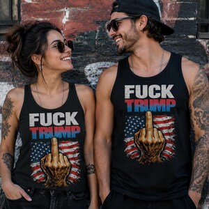 K&ouml;nnte beinhalten: Schwarze Tanktops mit dem Text "FUCK TRUMP" in wei&szlig;en und roten Buchstaben &uuml;ber einer Grafik der amerikanischen Flagge und einer Hand, die den Mittelfinger zeigt. Die Tanktops werden von zwei Personen getragen.