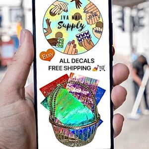May include: A phone screen displaying a logo for "JVA Nail Supply" with a circle of hands showing nail art designs. The text "All Decals Free Shipping" is below the logo. A basket filled with colorful holographic nail decals is shown below the text.