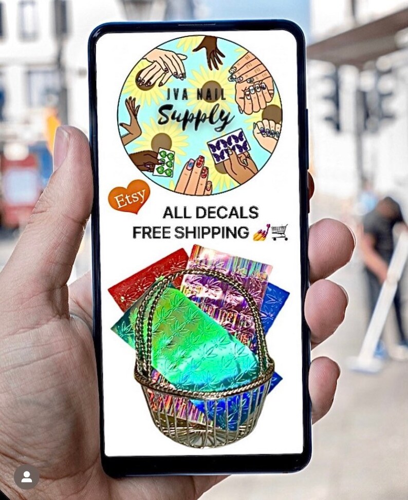 May include: A phone screen displaying a logo for "JVA Nail Supply" with a circle of hands showing nail art designs. The text "All Decals Free Shipping" is below the logo. A basket filled with colorful holographic nail decals is shown below the text.