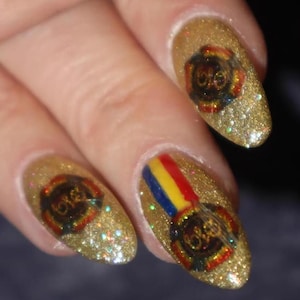 May include: Close-up of gold glitter nail polish with a design featuring a logo with the letters "ELO" in a circular emblem. One nail has a rainbow stripe design. The nails are on a hand.