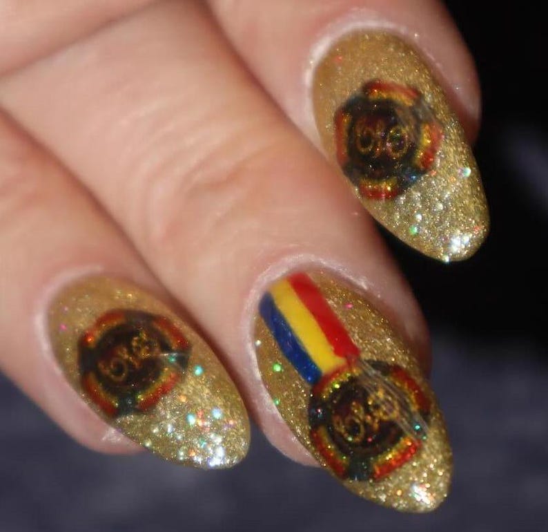 May include: Close-up of gold glitter nail polish with a design featuring a logo with the letters "ELO" in a circular emblem. One nail has a rainbow stripe design. The nails are on a hand.