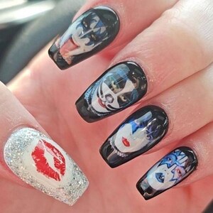 May include: Black acrylic nails with images of the band KISS. One nail features a red lip print on a silver glitter background. The other nails have images of the band members' faces.
