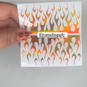 May include: Two sheets of iridescent flame nail decals. The decals are white with a holographic finish that shifts from orange to yellow to green. The decals are arranged in a repeating pattern of flames. The text "@jvanailsupply" is printed on the sheet.