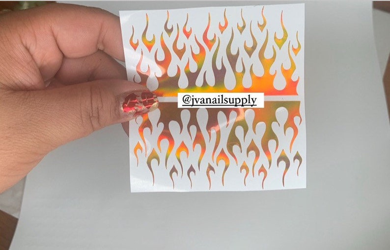 May include: Two sheets of iridescent flame nail decals. The decals are white with a holographic finish that shifts from orange to yellow to green. The decals are arranged in a repeating pattern of flames. The text "@jvanailsupply" is printed on the sheet.