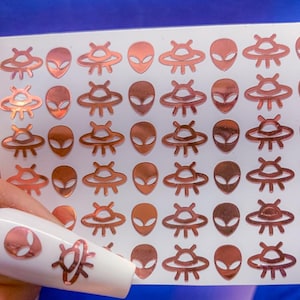 May include: A sheet of rose gold alien and UFO stickers. The stickers are arranged in a grid pattern on a white background.